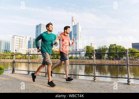 Photo of two strong sporty men wearing sportswear doing workout and running on city riverfront Stock Photo