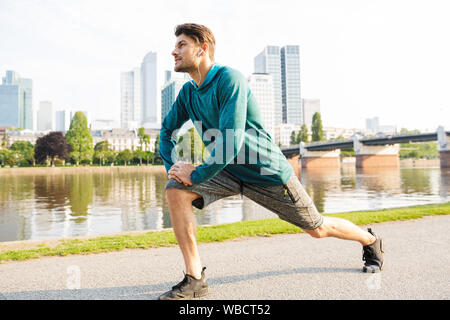 Image of attractive concentrated serious young strong sportsman outdoors in park listening music with earphones make exercises for legs. Stock Photo