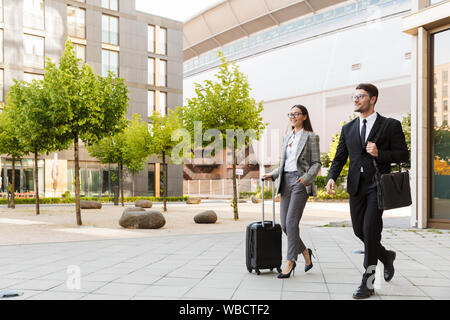 Two attractive smiling young office colleagues wearing suits walking outdoors at the city streets, carrying suitcase Stock Photo