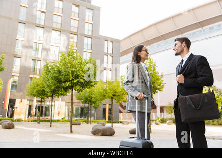 Two attractive smiling young office colleagues wearing suits standing outdoors at the city streets, carrying suitcase, talking Stock Photo