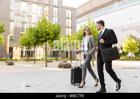 Two attractive smiling young office colleagues wearing suits walking outdoors at the city streets, carrying suitcase, talking Stock Photo