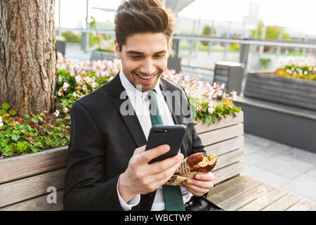 Attractive smiling young businessman wearing a suit sitting on a bench outdoors at the city street, having lunch break, using mobile phone Stock Photo