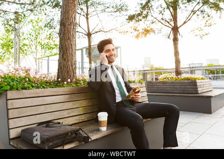 Attractive smiling young businessman wearing a suit sitting on a bench outdoors at the city street, having lunch break, talking on mobile phone Stock Photo