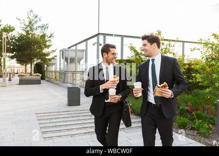 Two happy attractive young businessmen wearing suits walking outdoors at the city streets, having lunch Stock Photo