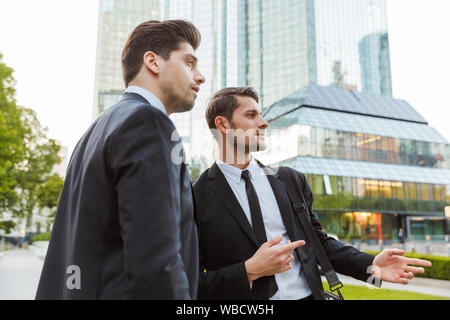Two attractive confident young businessmen wearing suits standing outdoors at the city streets, discussing new project Stock Photo