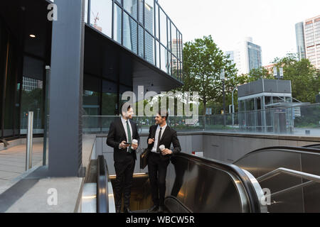 Two attractive confident young businessmen wearing suits walking outdoors at the city streets, having cup of coffee, moving on an escalator Stock Photo
