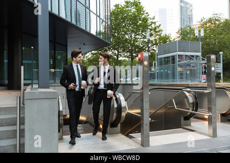 Two attractive confident young businessmen wearing suits walking outdoors at the city streets, having cup of coffee Stock Photo