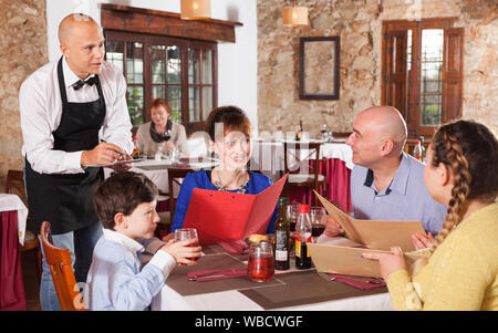 Waiter in white shirt and black apron taking order of family with child at restaurant Stock Photo