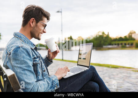 Side view of a handsome smiling young man dressed casually spending time outdoors at the city, using laptop computer while sitting on a bench and drin Stock Photo