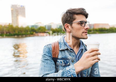 Handsome confident young man dressed casually spending time outdoors at the city, carrying backpack, holding takeaway cup Stock Photo