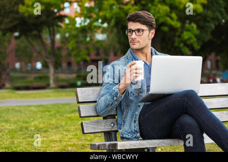 Handsome young man dressed casually spending time outdoors at the city, using laptop computer while sitting on a bench and drinking takeaway coffee Stock Photo