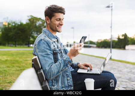 Handsome smiling young man dressed casually spending time outdoors at the city, using laptop computer while sitting on a bench and using mobile phone Stock Photo