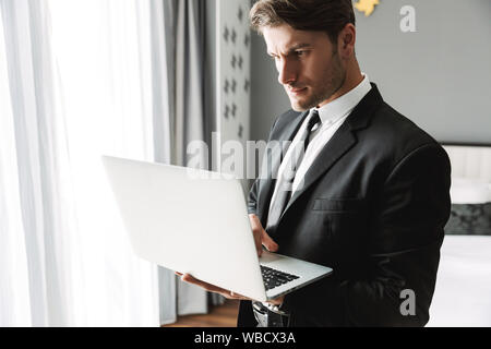 Image of concentrated young man wearing formal black suit standing by window with laptop computer in hotel apartment during business trip Stock Photo