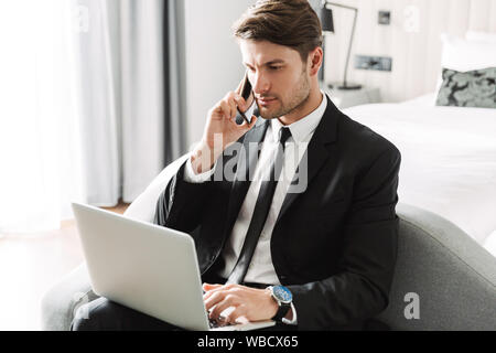 Image of modern young man wearing formal black suit talking on smartphone and using laptop computer in hotel apartment during business trip Stock Photo