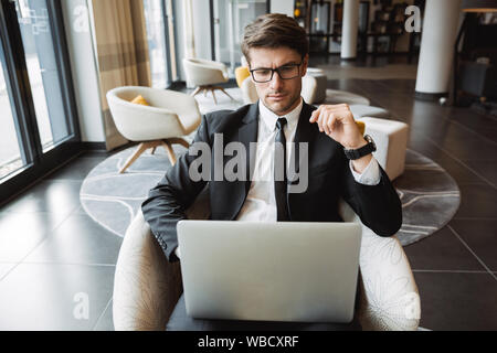 Photo of caucasian businessman wearing formal black suit and eyeglasses using laptop computer in hotel lobby Stock Photo