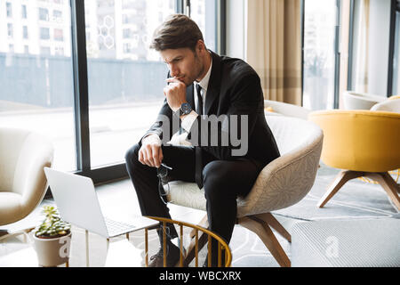Photo of concentrated businessman wearing formal black suit using laptop computer in hotel lobby Stock Photo