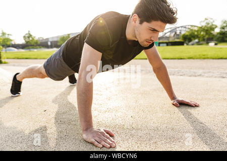 Image of muscular man in sportswear working out and doing push-ups at green park outdoors in morning Stock Photo