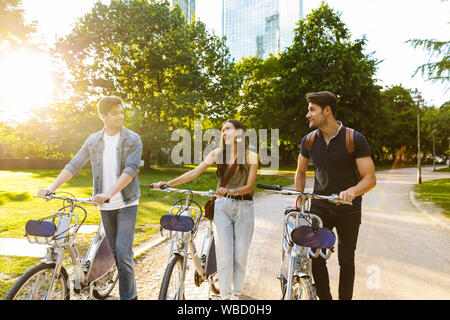 Photo of pleased young friends walking in nature green park outdoors on bicycles. Stock Photo