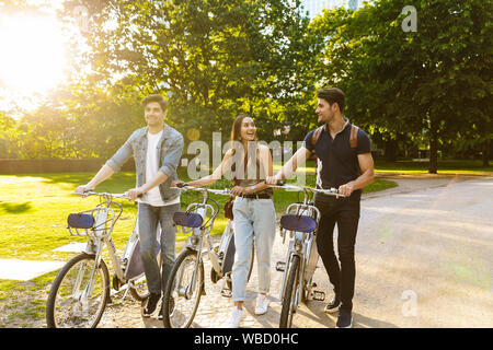 Photo of cheery happy young friends walking in nature green park outdoors on bicycles talking with each other. Stock Photo