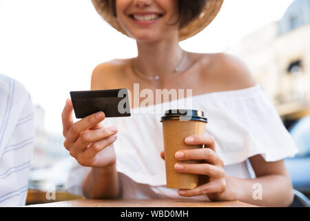 Cropped image of a cheerful young optimistic girl sitting outdoors in cafe drinking coffee holding credit card. Stock Photo