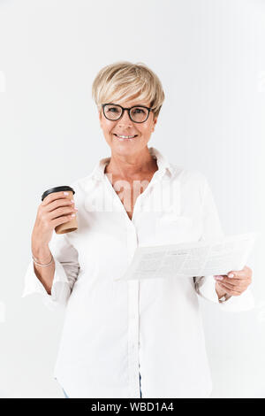 Portrait of happy adult woman wearing eyeglasses holding takeaway coffee cup and reading newspaper isolated over white background Stock Photo
