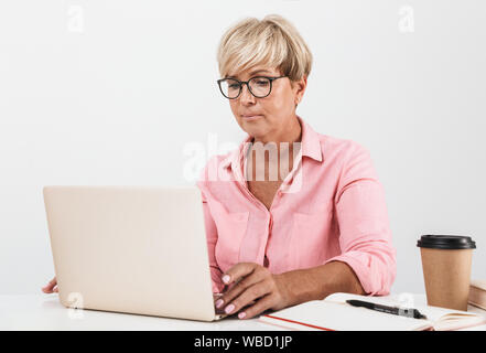 Portrait of elegant middle-aged woman with short blond hair wearing eyeglasses using laptop computer at table isolated over white background Stock Photo