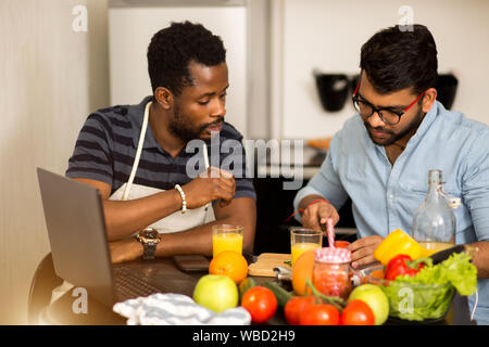 Two young men in casual clothes sitting at table, preparing salad, cutting vegetable, using laptop computer in kitchen in morning. Healthy food, cooki Stock Photo