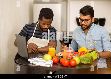 Shot of handsome bearded young indian man in glasses cutting vegetables on wooden chopping board while his serious african friend using mobile phone a Stock Photo