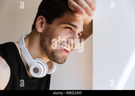 Photo of a cheerful smiling happy young sports man standing near window with headphones on neck. Stock Photo