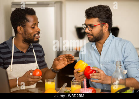 Closeup shot of two multi ethnic bearded friends in casual clothes preparing vegetable salad or smoothie, sitting at table talking using laptop comput Stock Photo
