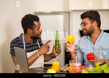 Handsome african american young man and his indian friend sitting at table holding vegetables, discussing recipe, looking at each other, using laptop Stock Photo