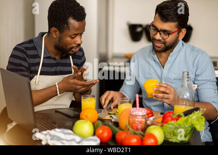Multi ethnic students sitting at table in kitchen watching video recipe on laptop and learn to cook healthy food. African guy in apron with his indian Stock Photo