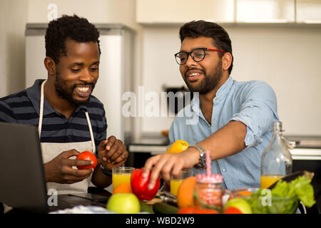Joyful multi ethnic students cooking and having fun in kitchen. African american guy and his indian friend sitting at table watching culinary TV show Stock Photo