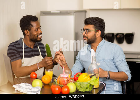 Two students argue what to cook for dinner. Multicultural friends sitting in kitchen, using laptop computer talking and preparing vegetable salad, smo Stock Photo
