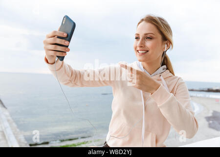 Image of smiling woman wearing tracksuit listening using earphones and pointing finger at cellphone near seaside in morning Stock Photo