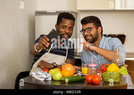Two multiracial friends students sitting at table taking selfie together while preparing vegetable salad and fruit juice in hostel. African guy with i Stock Photo
