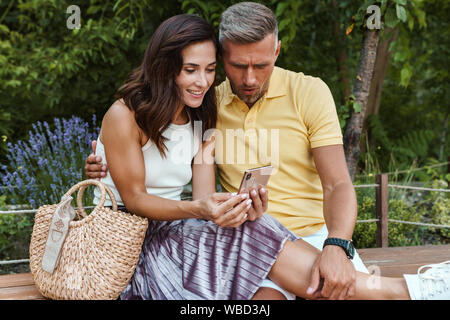 Portrait of surprised middle-aged couple man and woman looking at smartphone and hugging while sitting in summer park Stock Photo