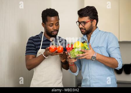 Handsome indian young man in glasses, blue t shirt holding bowl with salad leaves, bell peppers and talking with african american friend chef wearing Stock Photo