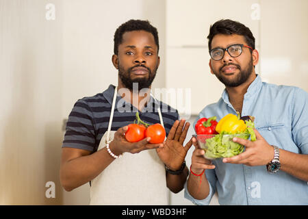 Closeup shot of two male chef friends standing in light kitchen and holding fresh organic vegetables and looking at camera. Healthy food, farm product Stock Photo