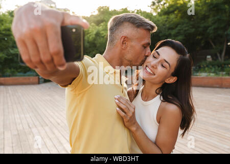 Portrait of pleased adult couple man and woman taking selfie photo on cellphone and kissing while walking in summer park Stock Photo