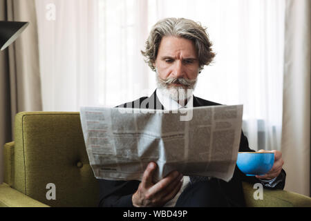 Photo of a mature handsome grey-haired businessman posing indoors at home dressed in formal clothes reading newspaper drinking coffee. Stock Photo