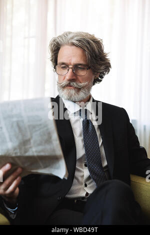 Image of a serious concentrated handsome grey-haired businessman wearing glasses posing indoors at home dressed in formal clothes reading newspaper. Stock Photo