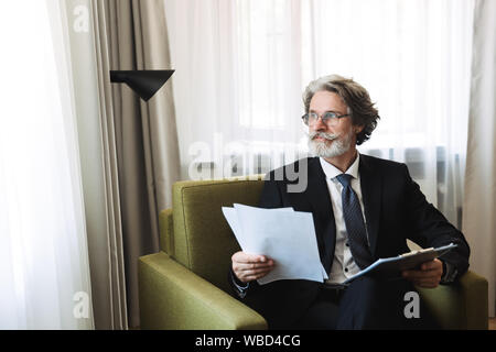 Portrait of pleased businessman wearing black suit and eyeglasses holding clipboard with paper documents while sitting on armchair in apartment Stock Photo