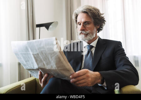 Photo of a serious grey-haired senior businessman indoors at home dressed in formal clothes reading newspaper. Stock Photo