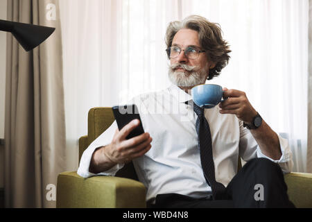 Photo of a concentrated thinking senior gray-haired business man sitting in chair indoors at home using smartphone drinking coffee. Stock Photo