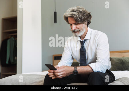 Photo of a concentrated senior gray-haired business man sitting on bed indoors at home using mobile phone. Stock Photo