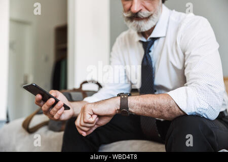 Cropped image of a concentrated senior gray-haired business man sitting on bed indoors at home using mobile phone looking at watch clock.