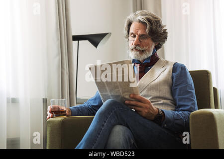 Image of businesslike adult professor man wearing stylish suit and eyeglasses reading newspaper while sitting on armchair in hotel apartment wih glass Stock Photo