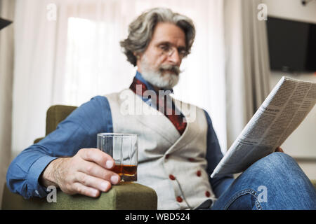 Image of successful adult professor man wearing stylish suit and eyeglasses reading newspaper while sitting on armchair in hotel apartment wih glass o Stock Photo