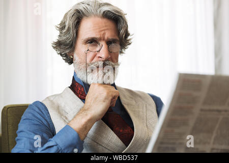 Image of smart adult professor man wearing stylish suit and eyeglasses reading newspaper while sitting on armchair in hotel apartment Stock Photo